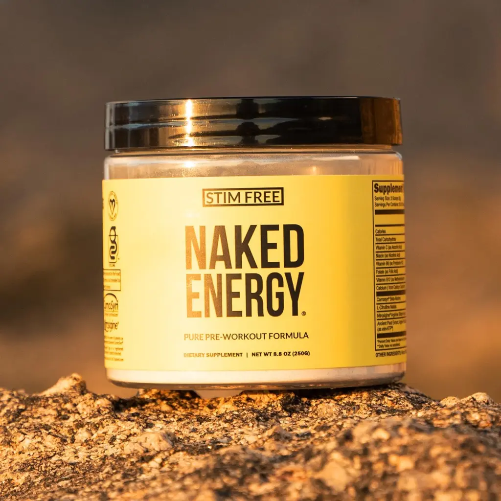 A jar of 'NAKED ENERGY' pre-workout supplement is positioned on a rocky surface. The label indicates it's a stimulant-free, pure pre-workout formula. The product is in a yellow container with black and white text, and the cap appears to be a dark color. The lighting suggests either an early morning or late afternoon setting, with warm tones casting across the scene.