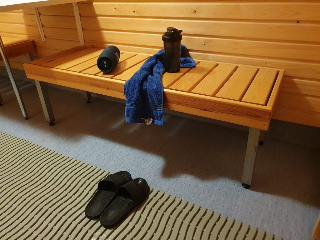 Speaker, water bottle, and towel on a wooden bench outside a sauna, with sandals on the floor.