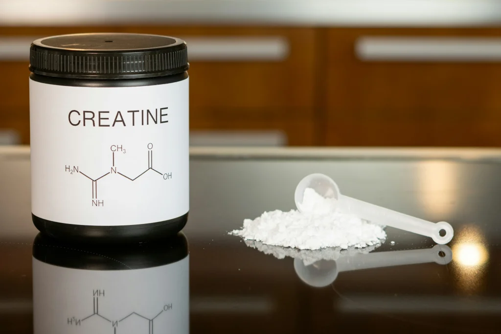 A black container labeled "CREATINE" with a chemical structure diagram displayed on the label, sitting on a reflective black surface. Next to it lies a white measuring scoop spilling a pile of white powder. The background is a kitchen with wooden cabinet doors.