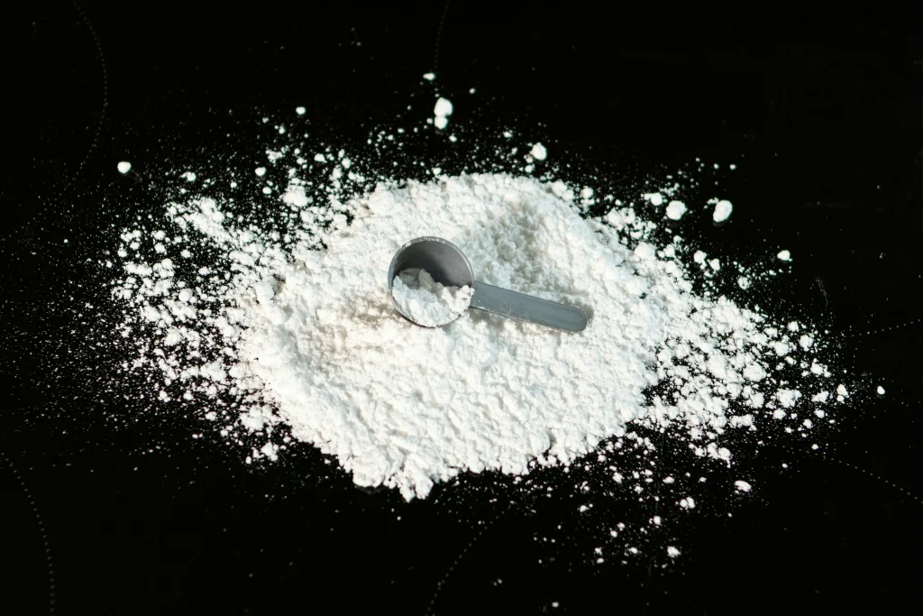 A close-up photograph of a gray scoop partially buried in white powder on a black background. The scattered powder and scoop are highly contrasted against the dark surface, with fine particles of the powder visible in the air, suggesting movement, possibly from the scoop being lifted. The image is detailed, showing the texture of the powder and the smoothness of the scoop's surface