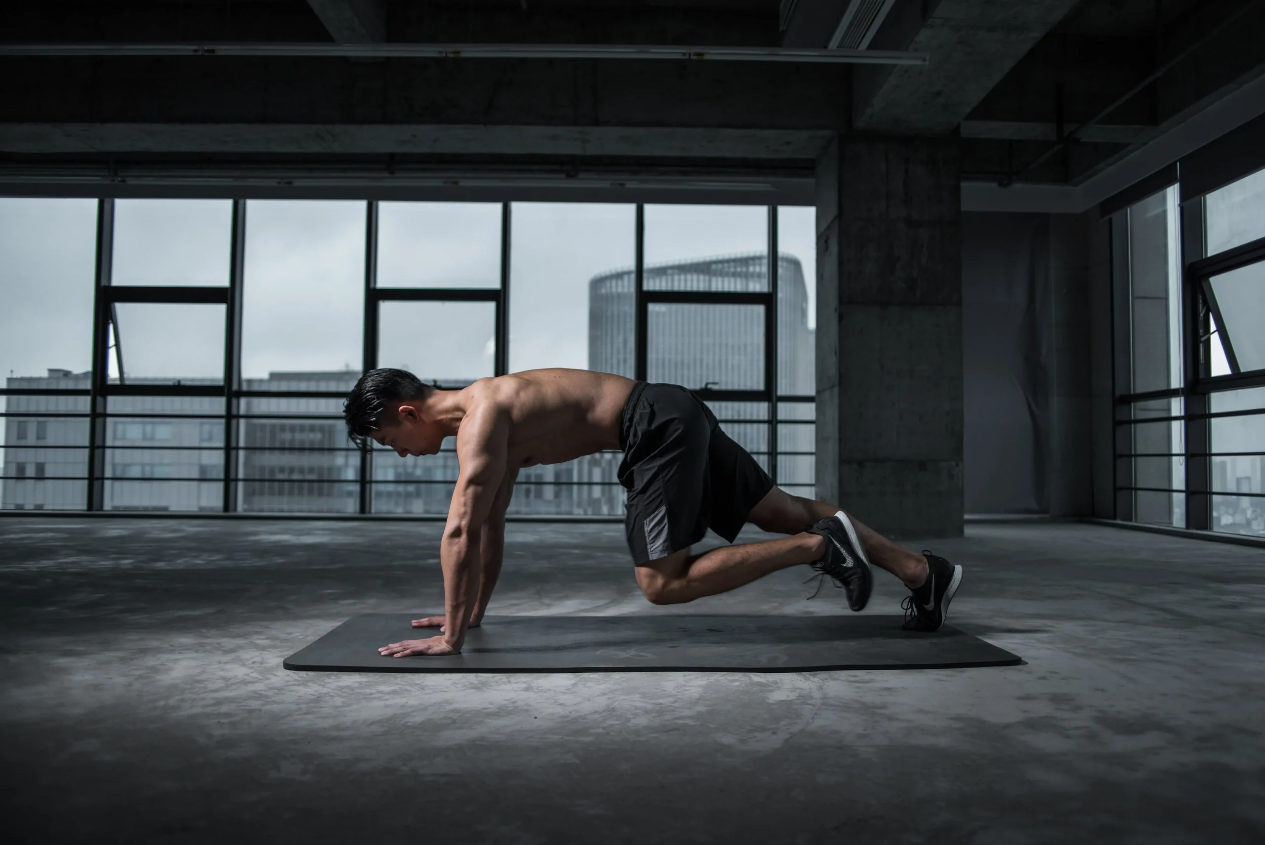 A muscular man, dressed in black shorts and no shirt, is performing mountain climbers on a yoga mat in an empty room.