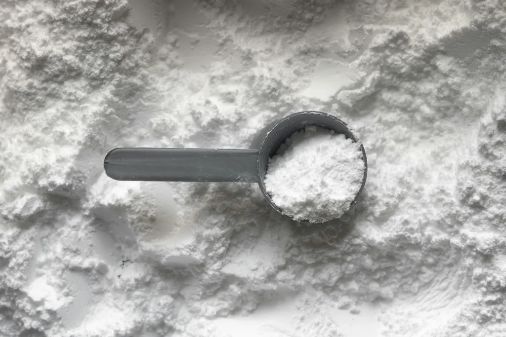 A close-up photograph showing a gray measuring scoop brimming with white creatine powder, questioning "Does creatine expire," as it rests on a surface layered with an abundance of the same substance, highlighting the texture and consistency of the powder.
