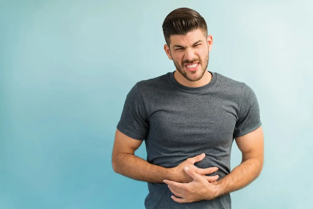 Young man wearing a gray t-shirt grimacing and holding his stomach as if in discomfort, standing against a light blue background – possibly experiencing digestive issues like those that can occur if one wonders, 'Does pre workout make you poop?