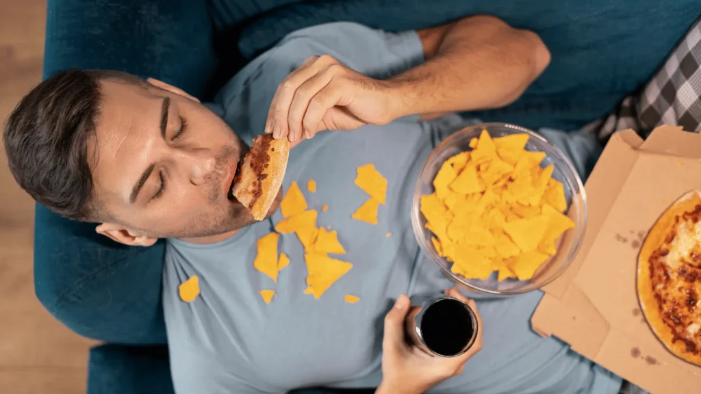 A man sits on a sofa, wondering if working out means he can eat pizza and chips, stuck in the " Can I Eat Whatever I Want If I Workout? " dilemma.