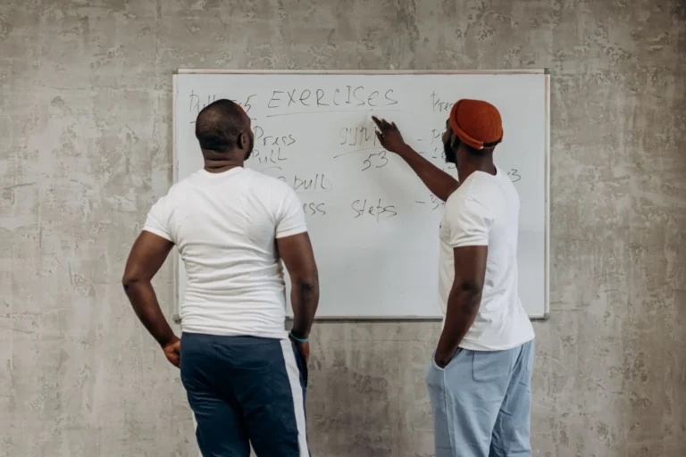 Two men planning a 6 day gym workout schedule on a whiteboard