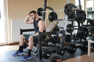 Man sitting thoughtfully beside a loaded weight bench, reflecting on a stalled workout plateau