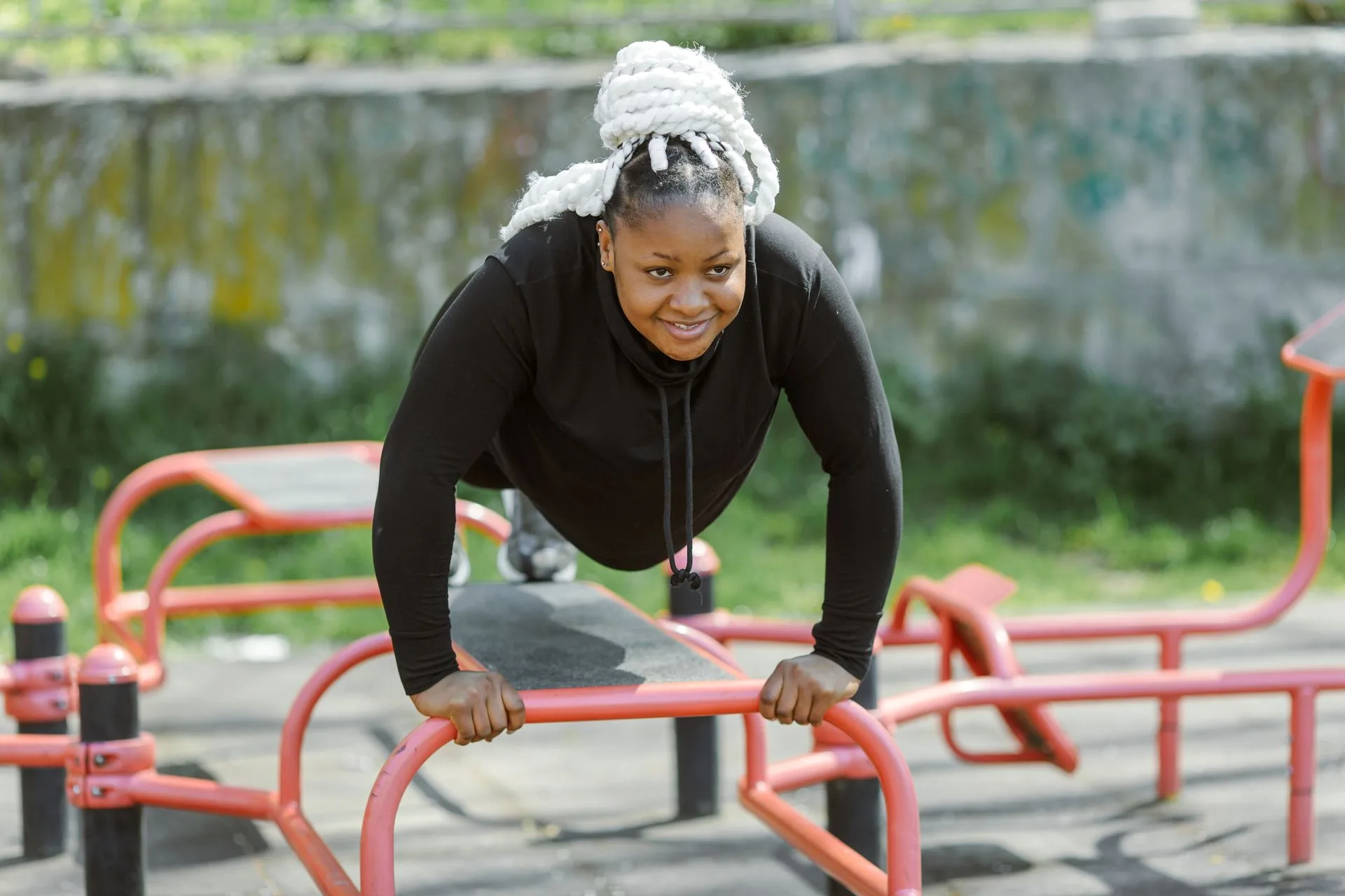 A smiling woman in black workout clothes doing incline push-ups on outdoor gym equipment in a park