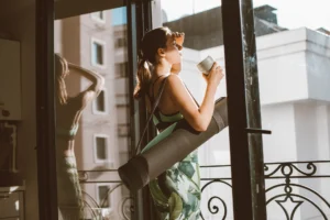 Woman in workout clothes with a yoga mat drinking coffee by a sunny balcony window