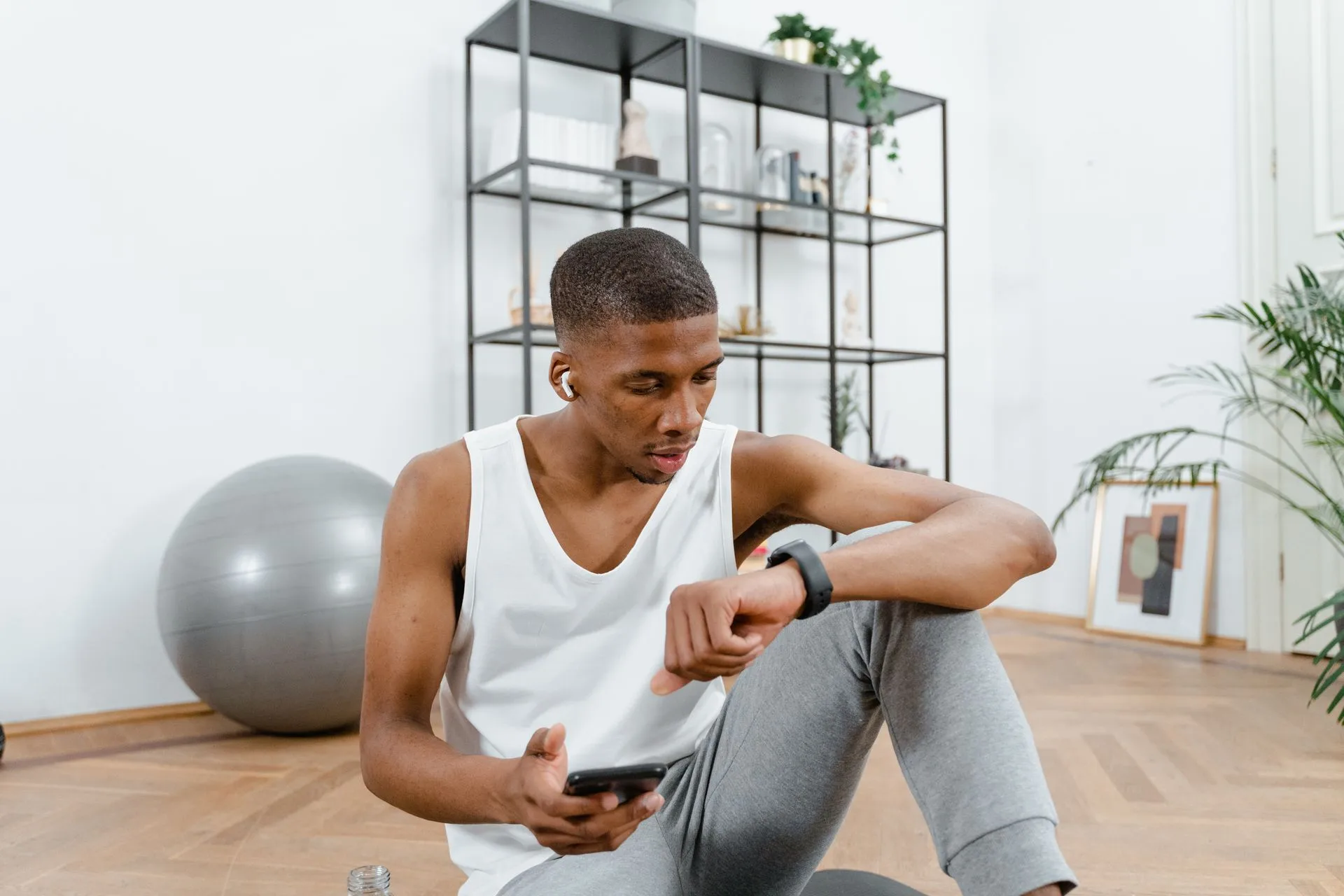 Man in a white tank top checking his smartwatch during a home workout